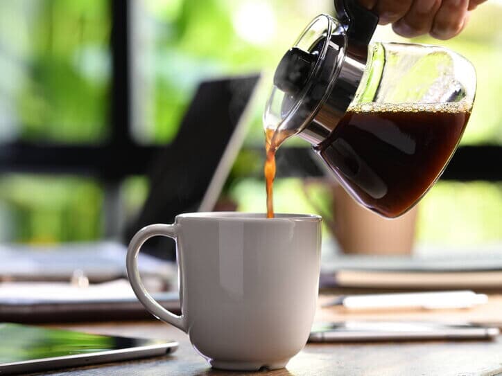 Eine Hand gießt frisch gebrühten Filterkaffee aus einer Kaffeekanne in eine weiße Tasse. Der Kaffee dampft, während auf dem Tisch ein Laptop und ein Tablet sichtbar sind. Der Hintergrund zeigt grüne Pflanzen.