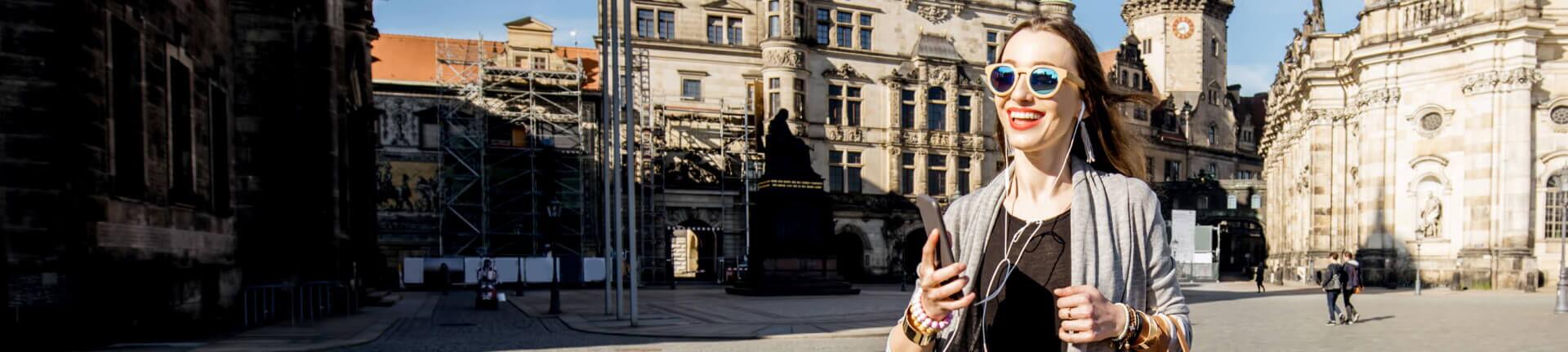 Eine Frau geht durch die Straßen von Dresden, vor der Kulisse historischer Gebäude. Sie trägt eine graue Strickjacke, schwarze Sonnenbrille, hört Musik über Kopfhörer und hält ihr Smartphone in der Hand.