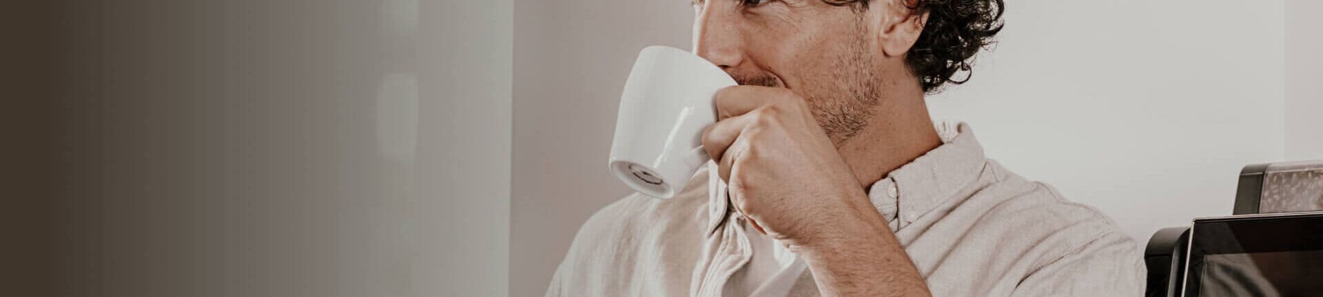 Ein Mann mit lockigem Haar genießt eine Tasse Kaffee, während er neben einem Kaffeevollautomaten von Kaffee Partner steht.